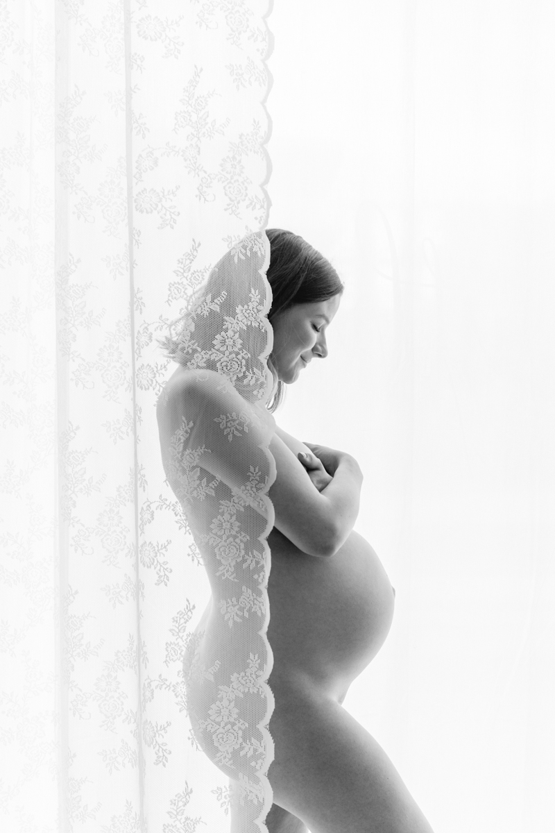 Schwanger Frau vor einem Fenster, leicht bedeckt mit weissem Spitzenvorhang