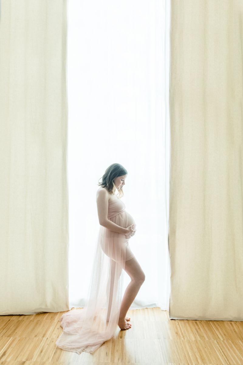 Schwangere Frau steht vor Fenster zwischen beigen Vorhängen. Sie trägt ein rosa Kleid und hält liebevoll ihren Babybauch
